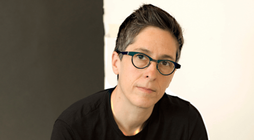 White person in black t-shirt and black glasses sits cross-legged against a black and white wall and looks at the camera