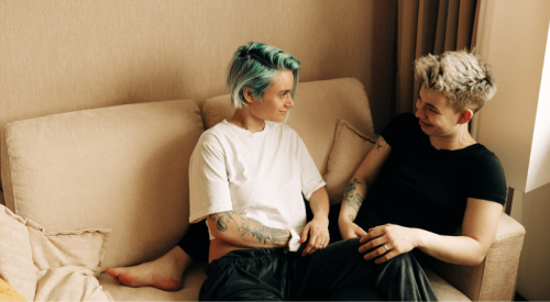 Two people, one with green hair and the other with bleached-blonde hair, sit in a beige living room on a sofa. They look at each other, and slightly smile.