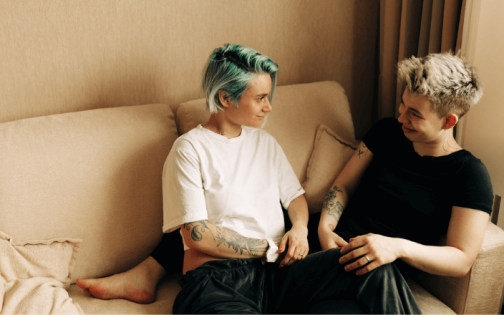 Two people, one with green hair and the other with bleached-blonde hair, sit in a beige living room on a sofa. They look at each other, and slightly smile.