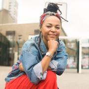 A Black woman on one knee in a concrete playground. She looks at the camera, has a hand propped beneath her chin, and wears a denim jacket and red trousers. Her hair is worn up, and a bandanna is tied around her head.