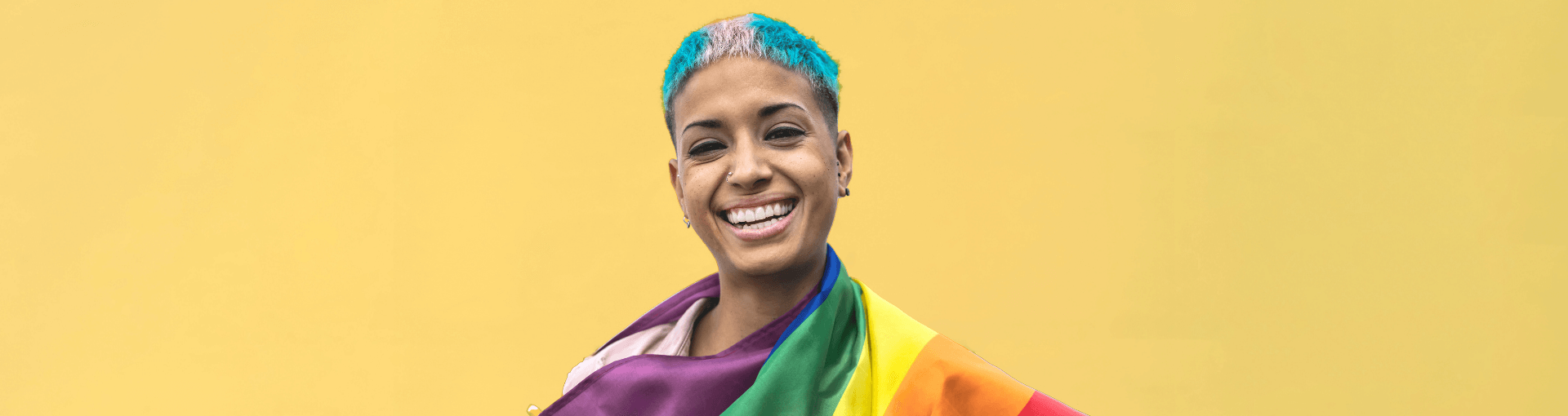 Person of colour with hair dyed in trans flag colours, draped in the rainbow flag, smiles at the camera